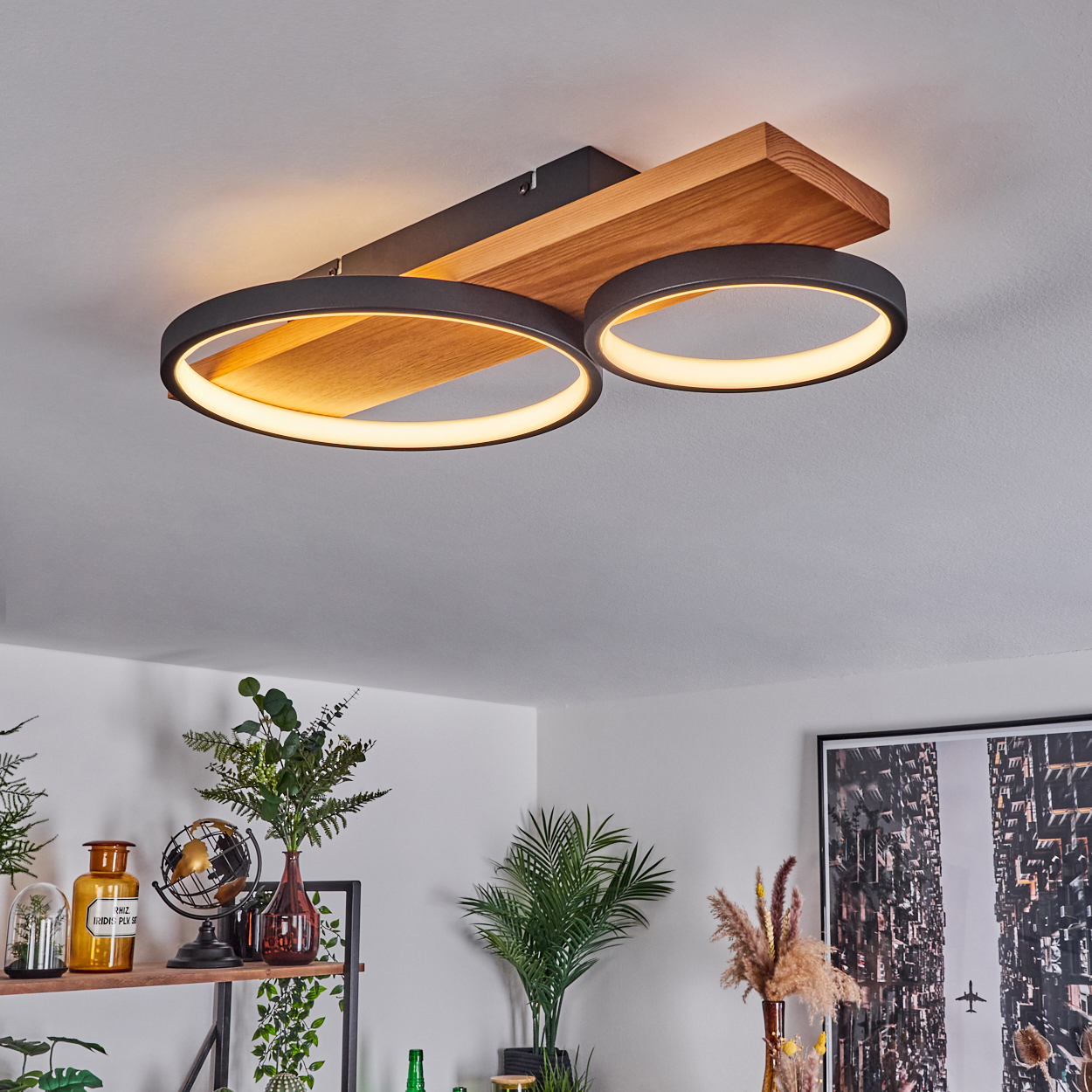 Moderne Deckenleuchte mit zwei kreisförmigen Leuchtelementen. Holz- und schwarze Metallstruktur. Innenraum mit Pflanzen und Dekoration.