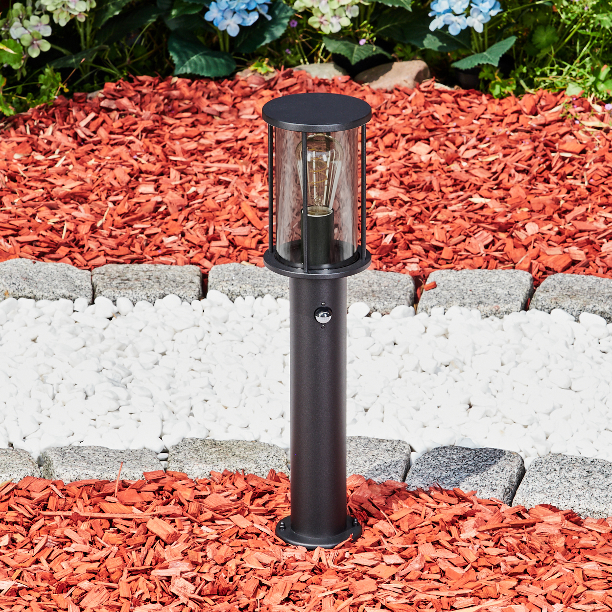 Eine schwarze Außenlampe steht auf roter Mulch, begrenzt von weißen Steinen und grauen Pflastersteinen. Blumen sind im Hintergrund.