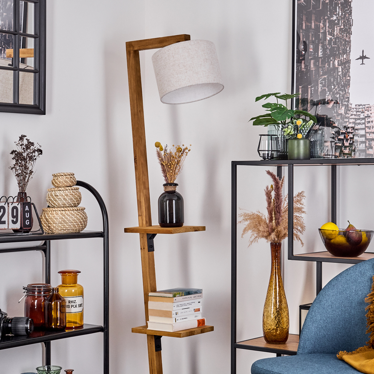 Eine Holzlampe und Regale zeigen Dekorationen. Ein blauer Stuhl und ein Stadtbildposter sind im Hintergrund. Verschiedene Objekte werden ausgestellt.
