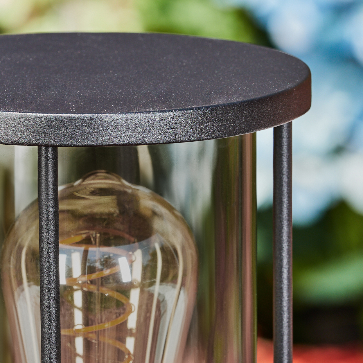 Nahaufnahme einer schwarzen Außenlampe mit Glaszylinder, Vintage-Glühbirne und schwarzen Metallstäben. Der Hintergrund ist unscharf.
