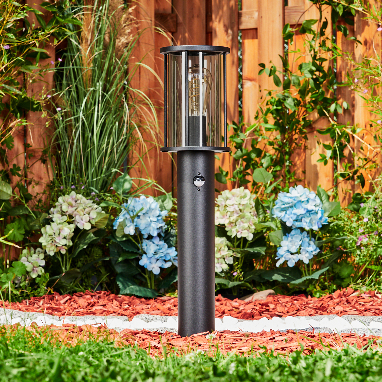 Eine schwarze Außenlampe steht in einem Garten mit Blumen und einem Holzzaun im Hintergrund.