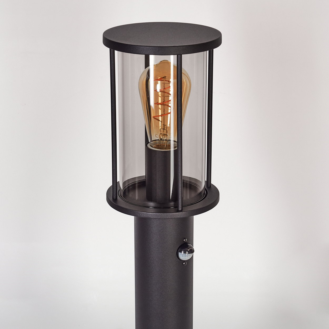 Eine schwarze Außenlampe mit einem Gehäuse aus klarem Glas. Im Inneren befindet sich eine Glühbirne im Vintage-Stil. Am Pfosten befindet sich ein Bewegungsmelder.
