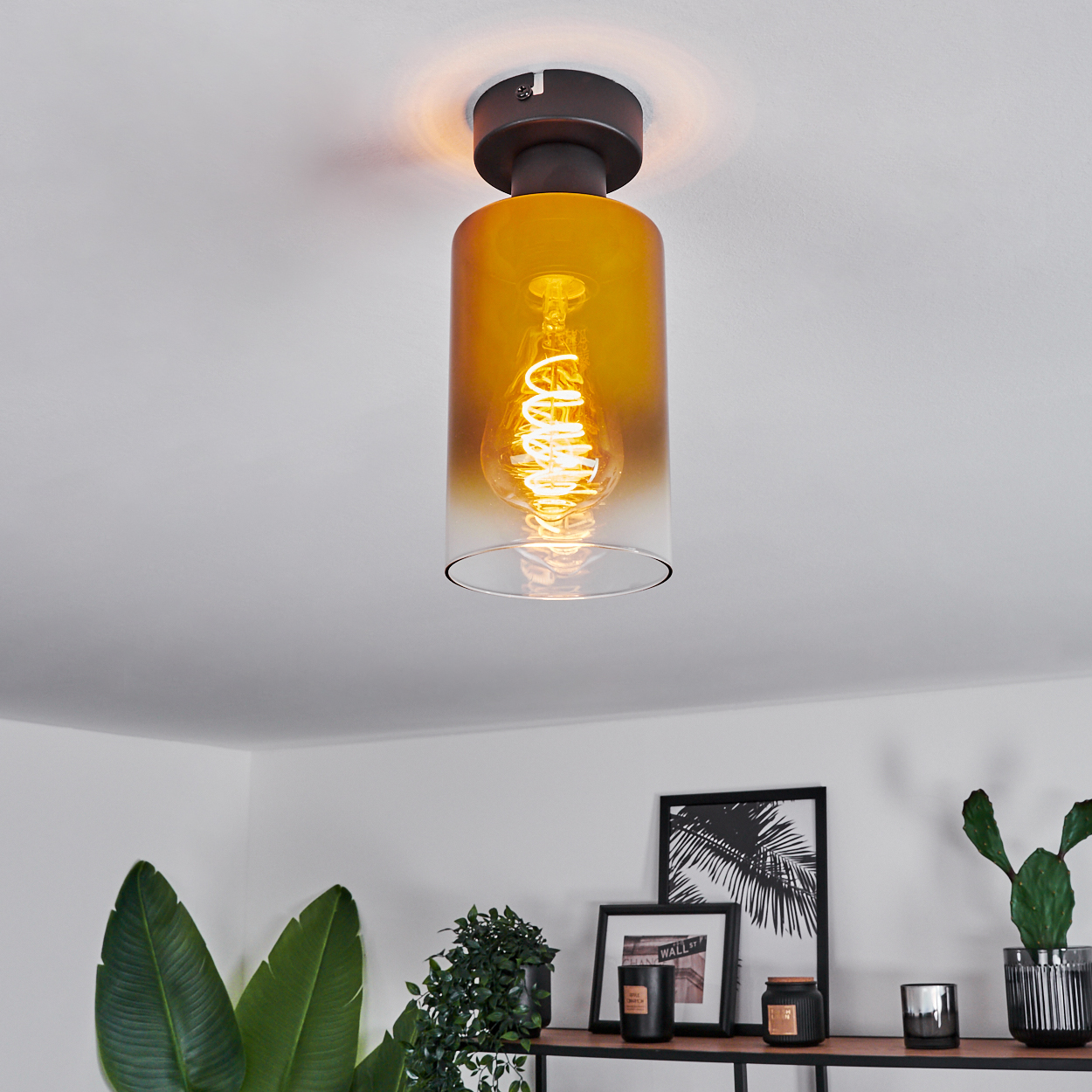 Eine Deckenlampe mit gelbem Glasschirm und sichtbarer Wendelgl ühbirne. Darunter ein Regal mit Pflanzen, Kerzen und gerahmten Bildern.