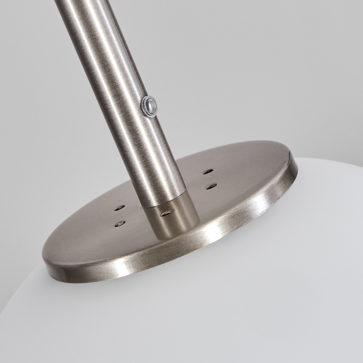 Nahaufnahme des Lampenfußes. Silberfarbener Metallpfosten an runder Silberbasis mit kleinen Löchern befestigt. Weiße Glaskugel darunter.