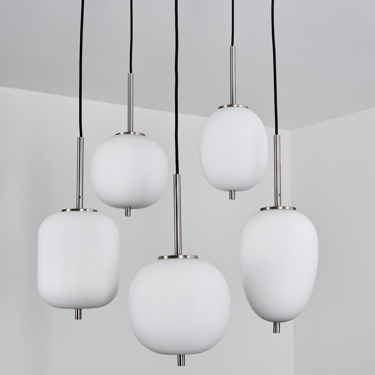 Fünf weiße Glas-Pendelleuchten hängen an schwarzen Schnüren. Jede hat einen silbernen Stiel und eine einzigartige abgerundete Form vor einer weißen Wand.