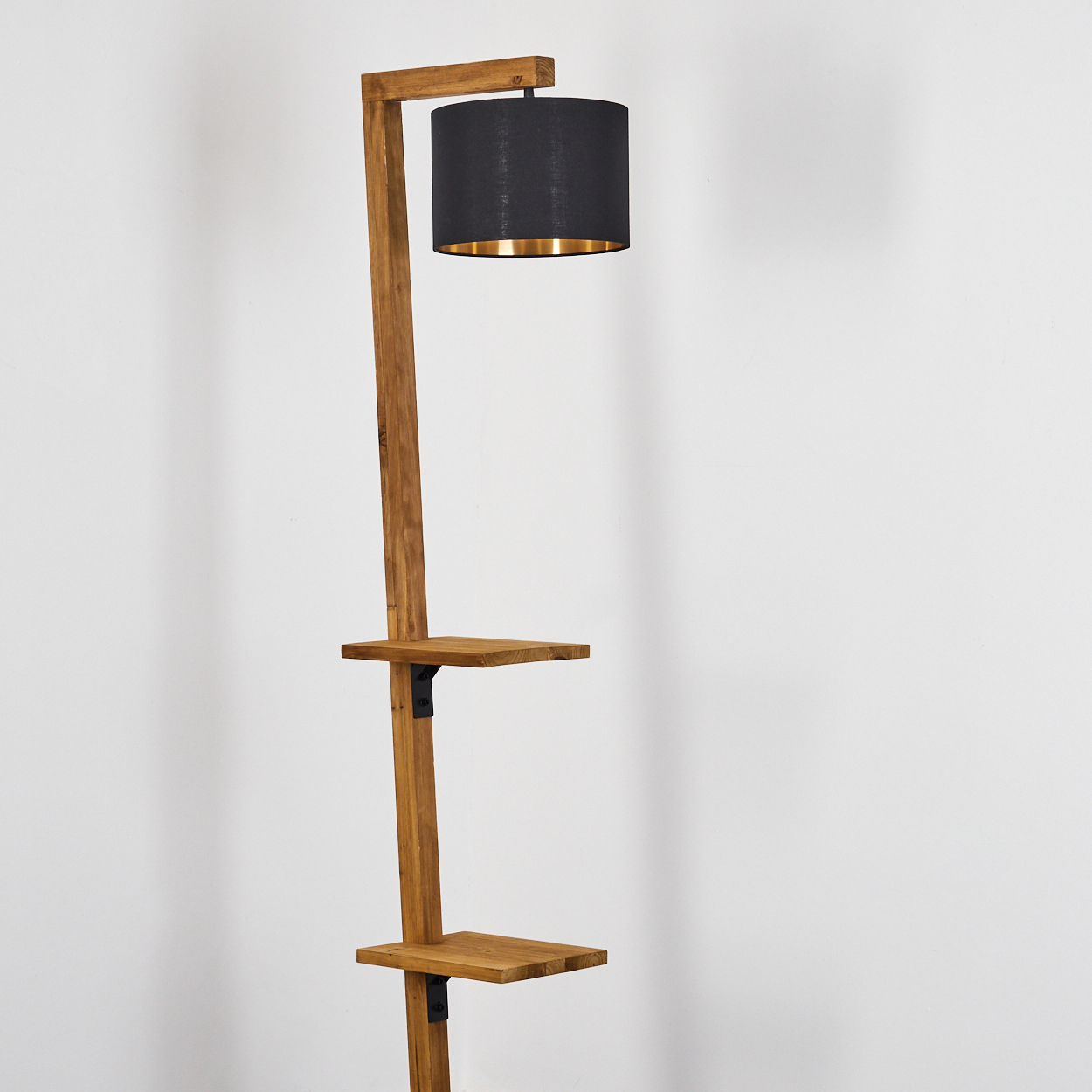 Eine Stehlampe aus Holz mit schwarzem Lampenschirm und goldenem Innenraum. Sie hat zwei Ablagen und steht vor einer weißen Wand.