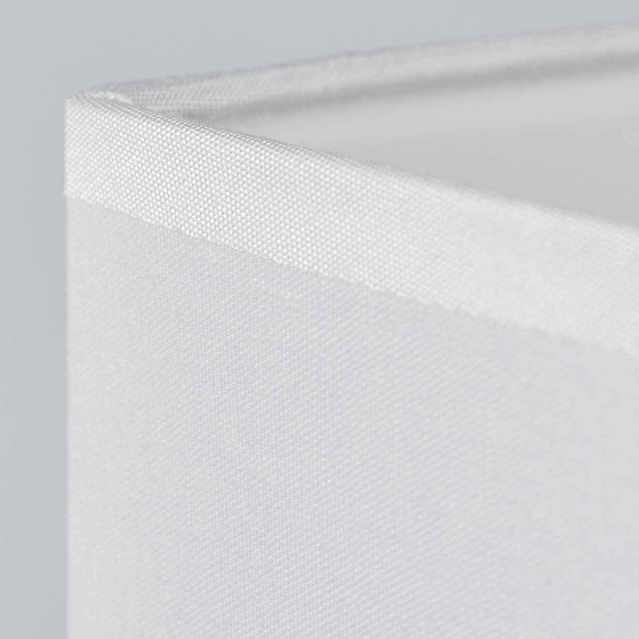 Nahaufnahme eines weißen, rechteckigen Lampenschirms. Der Stoff hat eine feine Textur. Die obere Kante hat eine etwas dunklere Einfassung.