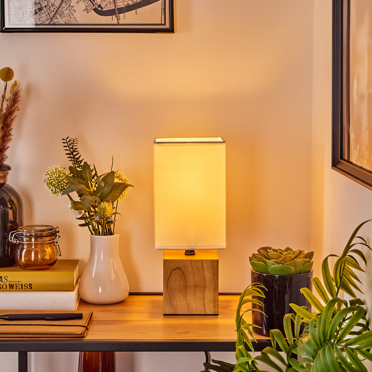 Eine beleuchtete Lampe steht auf einem Holztisch. Blumen, Bücher und Pflanzen umgeben sie. Eine Karte und ein gerahmtes Bild sind an der Wand.