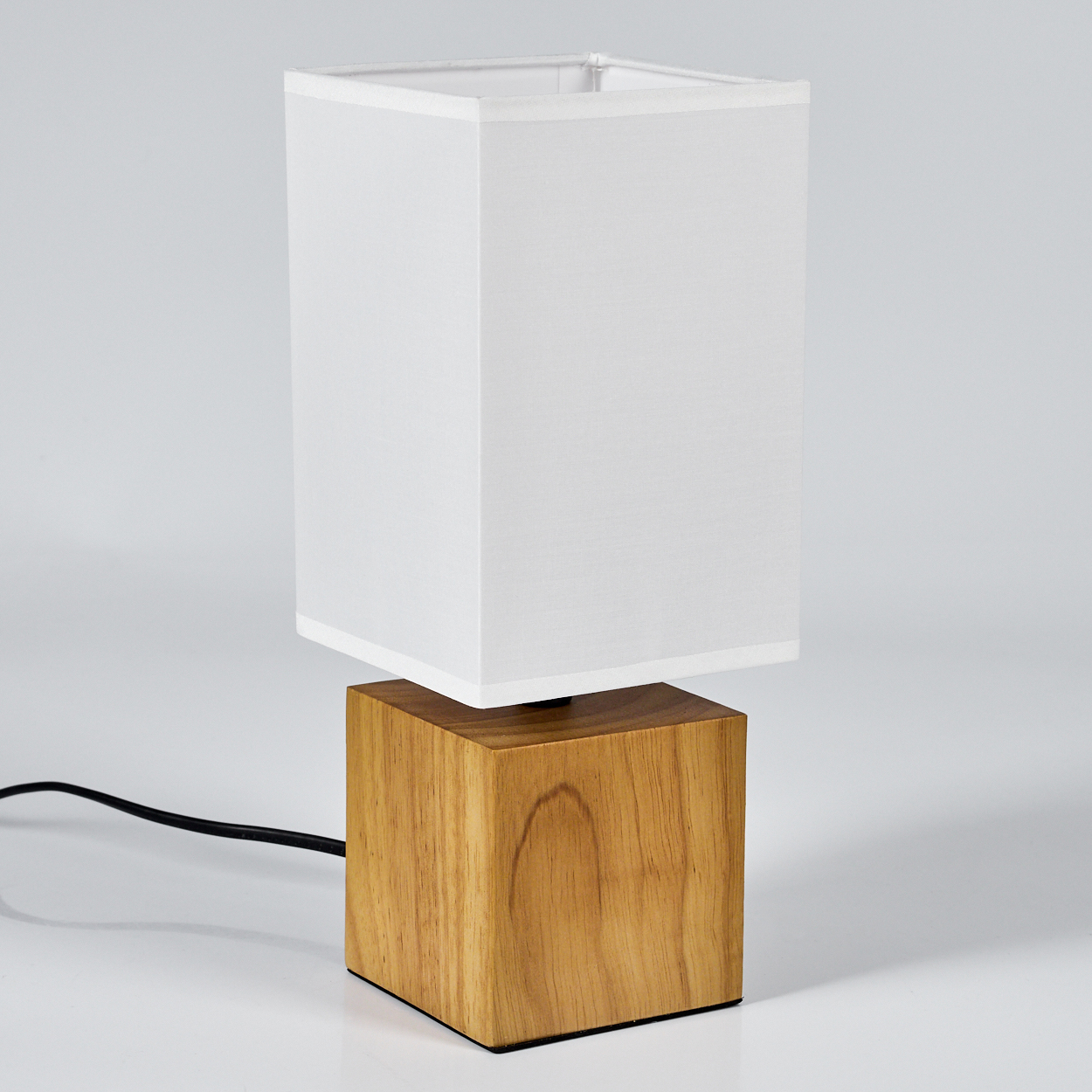 Eine Lampe mit weißem, rechteckigem Schirm auf einem quadratischen Holzsockel. Die Lampe steht auf einer weißen Oberfläche mit schwarzem Kabel.