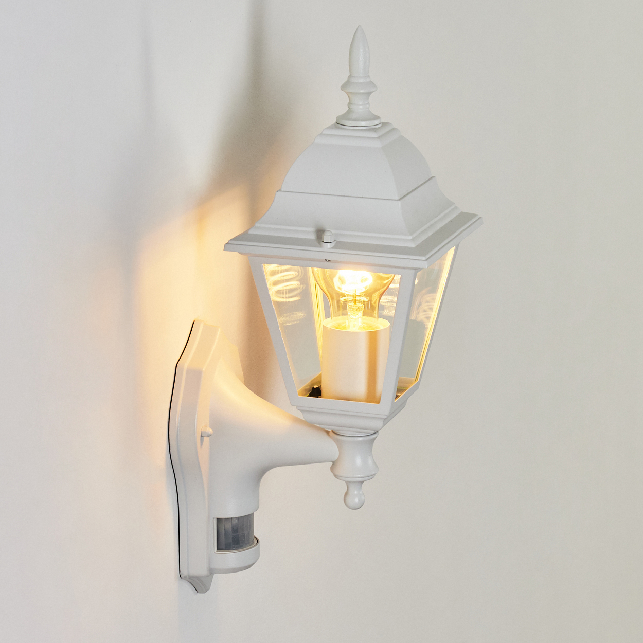 Eine weiße Außenlampe ist an einer weißen Wand befestigt. Die Lampe ist beleuchtet, mit einem Lichtsensor und einer Glühbirne.