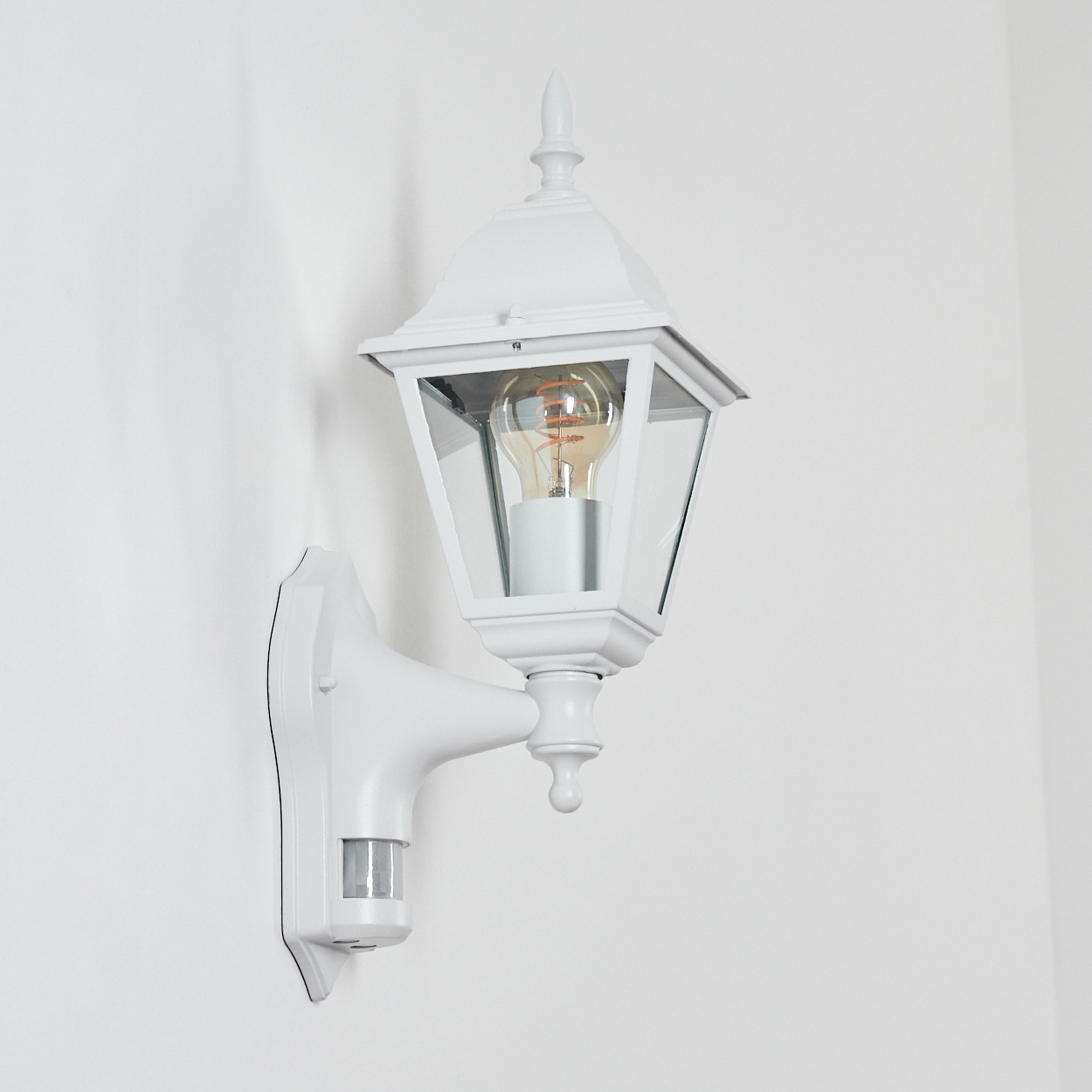 Eine weiße Außenlampe mit Glühbirne ist an einer weißen Wand befestigt. Sie hat einen Bewegungsmelder.