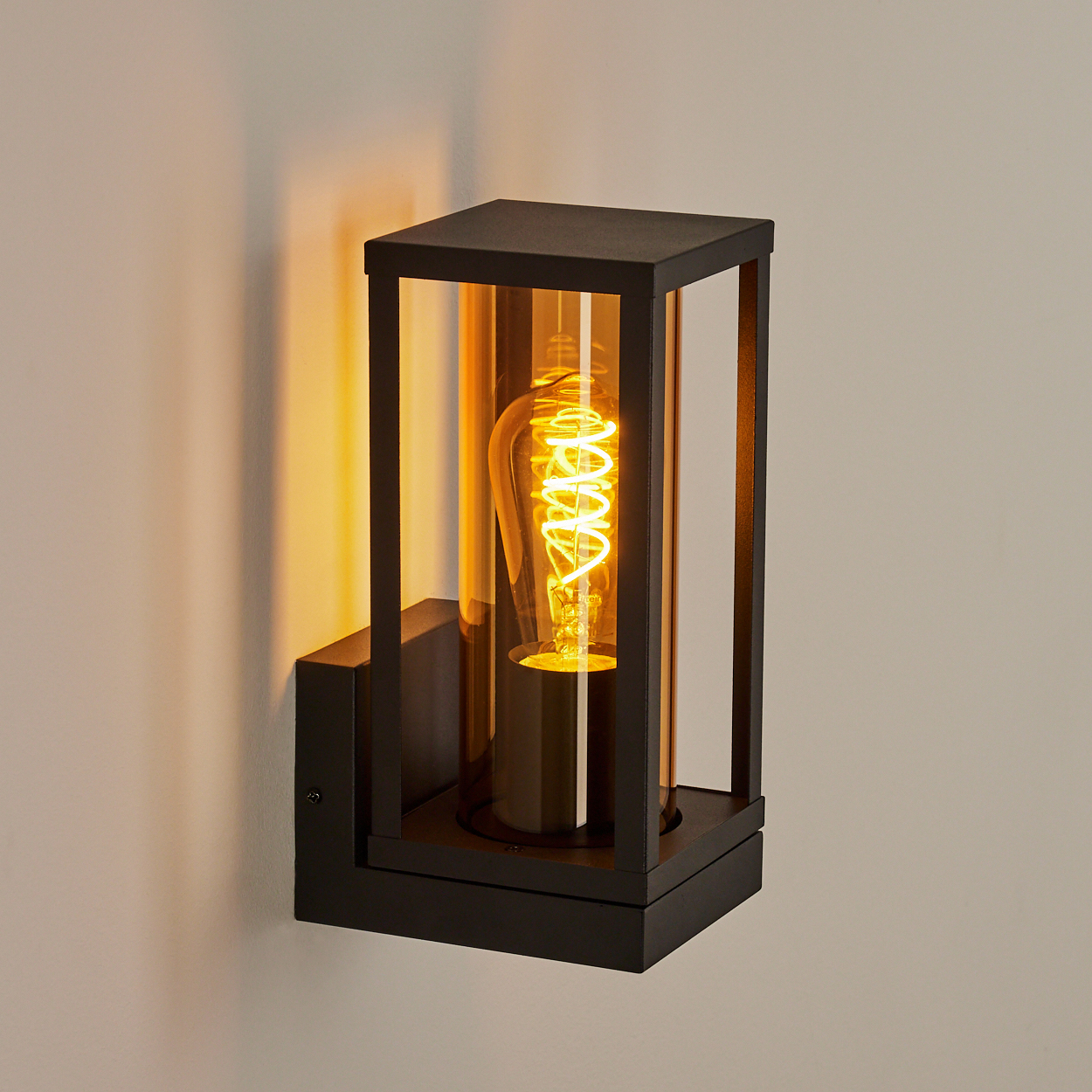 Eine schwarze Außenwandleuchte mit einer leuchtenden Wendelfadenbirne in einem Glaskasten. Die Lampe ist an einer hellen Wand montiert.