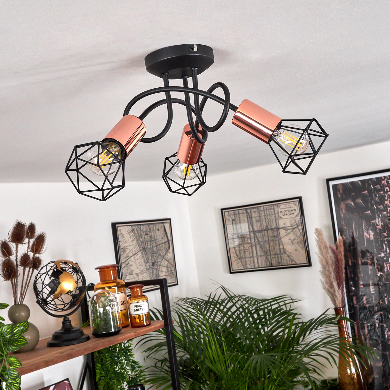 Deckenleuchte mit drei kupfer-schwarzen Leuchten. Im Hintergrund ein Regal mit Deko und Pflanzen sowie gerahmte Karten an der Wand.