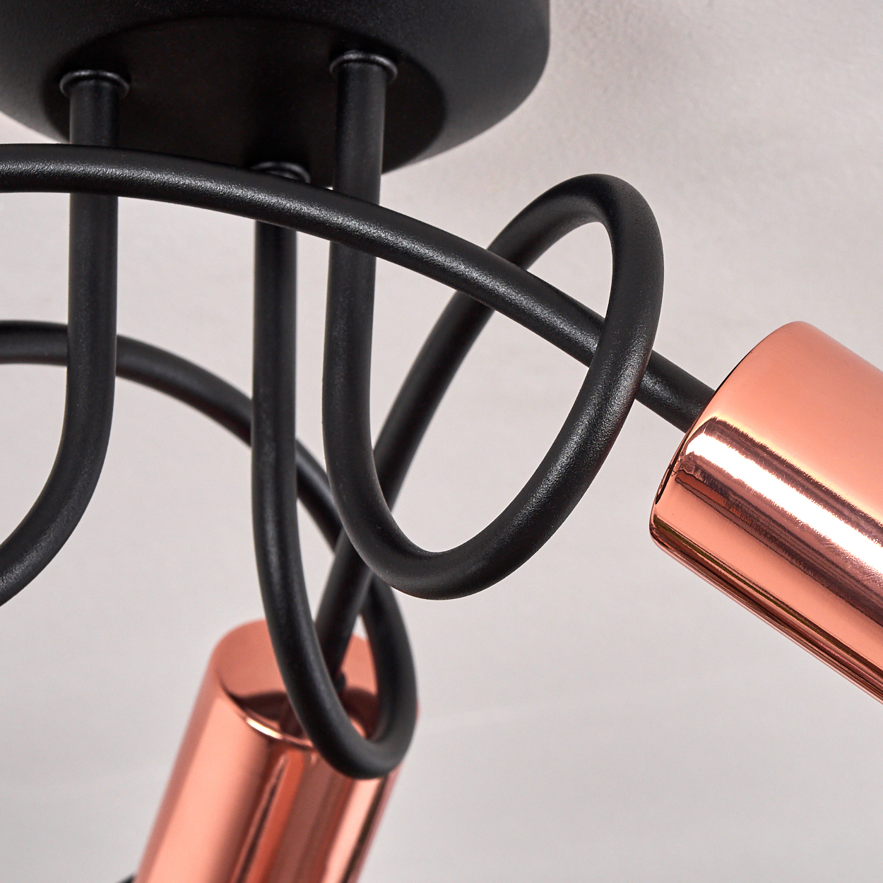 Nahaufnahme einer modernen schwarzen Deckenlampe mit kupferfarbenen zylindrischen Lampenschirmen. Der Hintergrund ist eine schlichte weiße Wand.