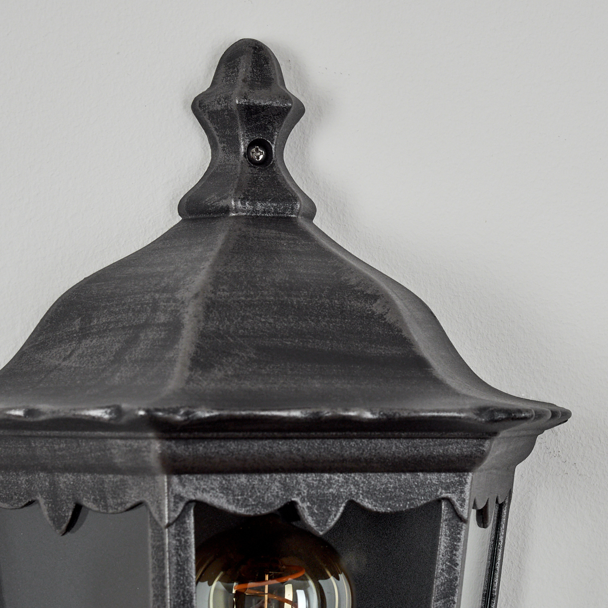 Nahaufnahme einer dunklen Metall-Außenlampe, montiert an einer hellgrauen Wand. Die Lampe hat eine dekorative Spitze und eine sichtbare Glühbirne.