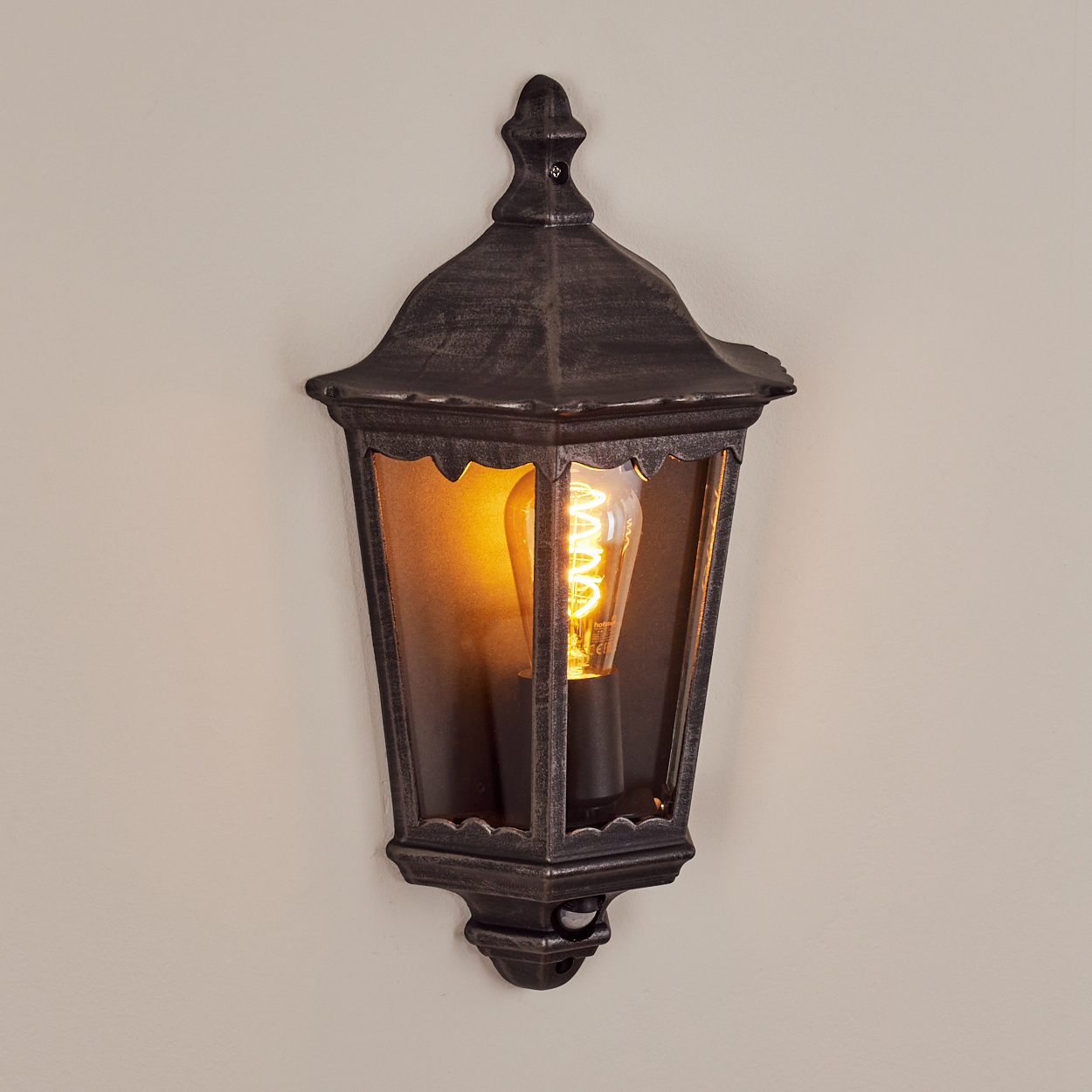 Eine Wandleuchte im antiken Stil mit einer glühenden Glühbirne im Inneren. Sie ist an einer beigefarbenen Wand montiert. Die Lampe ist dunkelgrau mit einem dekorativen Design.