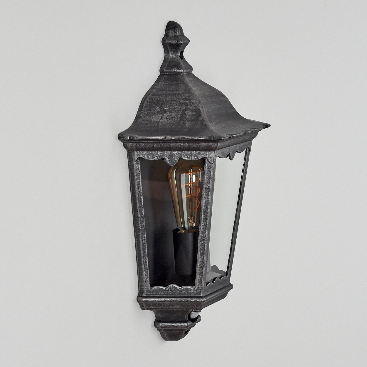 Eine dekorative, verzierte Wandlampe aus schwarzem Metall mit Glasscheiben. Im Inneren leuchtet eine Glühbirne im Vintage-Stil vor einer weißen Wand.