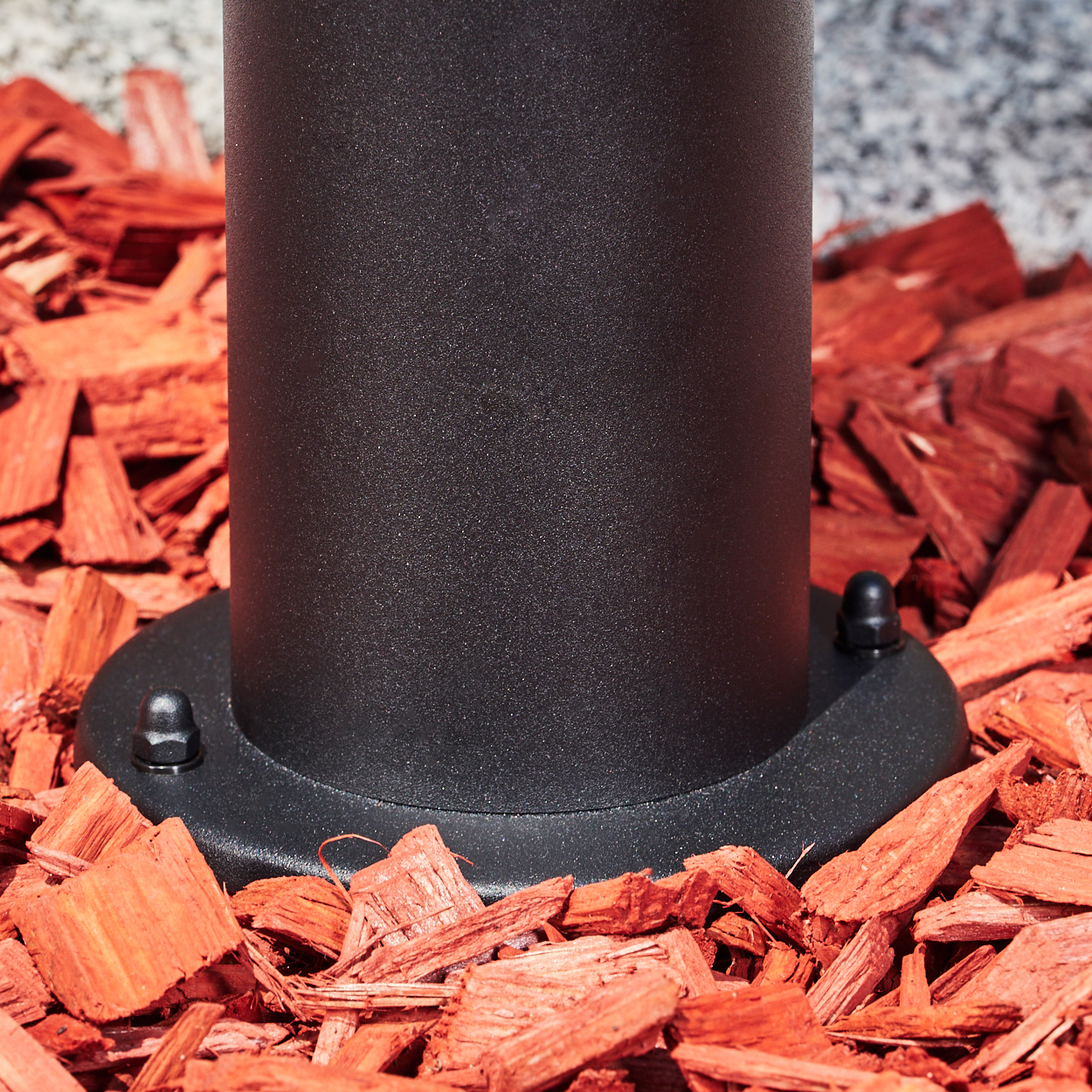 Nahaufnahme eines schwarzen Metallpfostenfußes, umgeben von roten Holzschnitzeln. Der Hintergrund hat eine Steinstruktur.