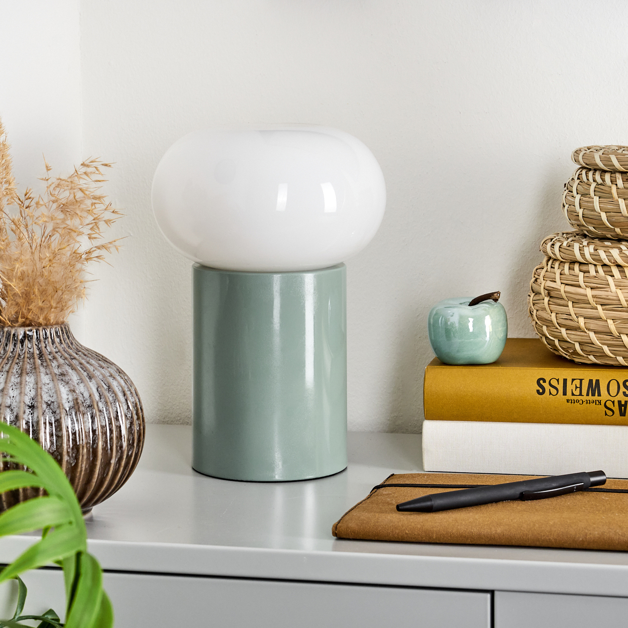 Ein Tisch mit einer weißen Lampe, einer Vase, Büchern, einem Stift und einem Notizbuch. Im Hintergrund sind Weidenkörbe.