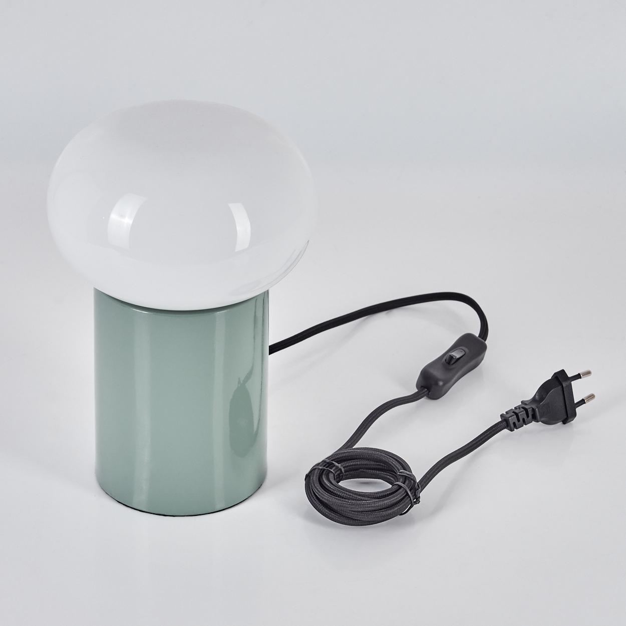 Eine pilzförmige Lampe mit weißem Schirm und grünem Sockel. Sie hat ein schwarzes Kabel mit Schalter und Stecker.