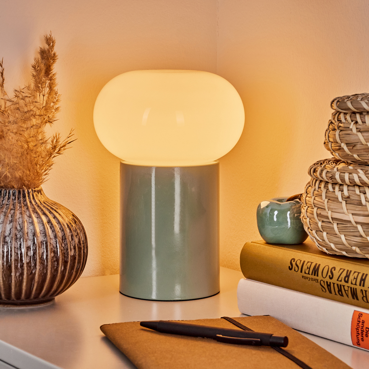 Eine Lampe mit grünem Sockel und weißem Licht steht auf einem weißen Tisch. Daneben Bücher, eine kleine Vase und ein Stift.