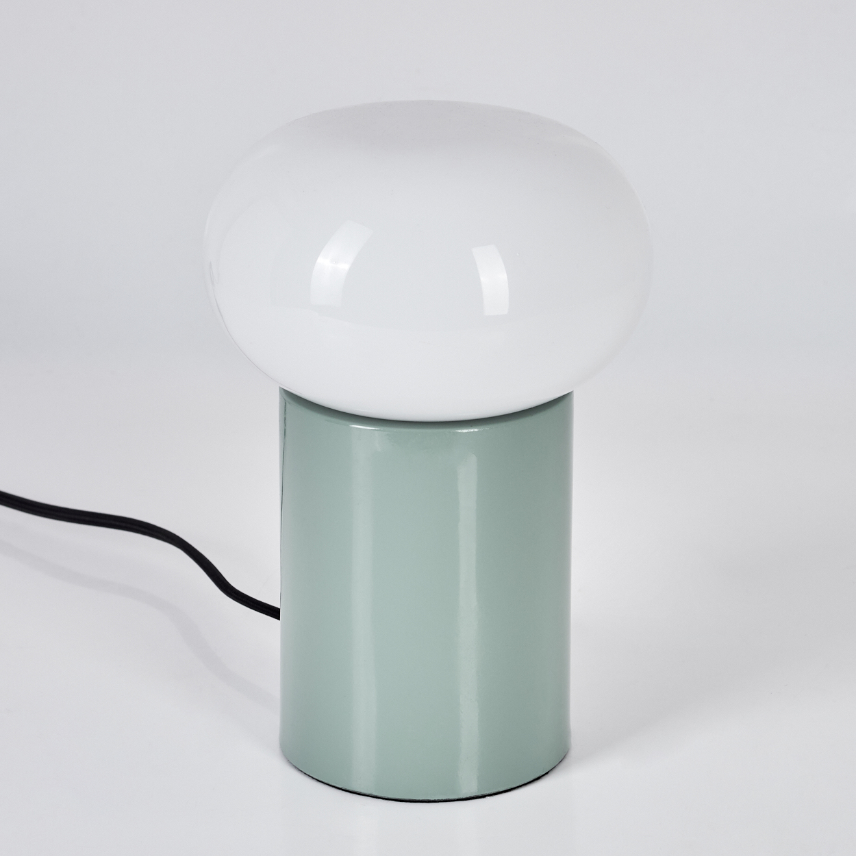 Eine moderne Lampe mit weißem, ovalem Lampenschirm auf einem hellgrünen zylindrischen Sockel. Ein schwarzes Netzkabel ist sichtbar.