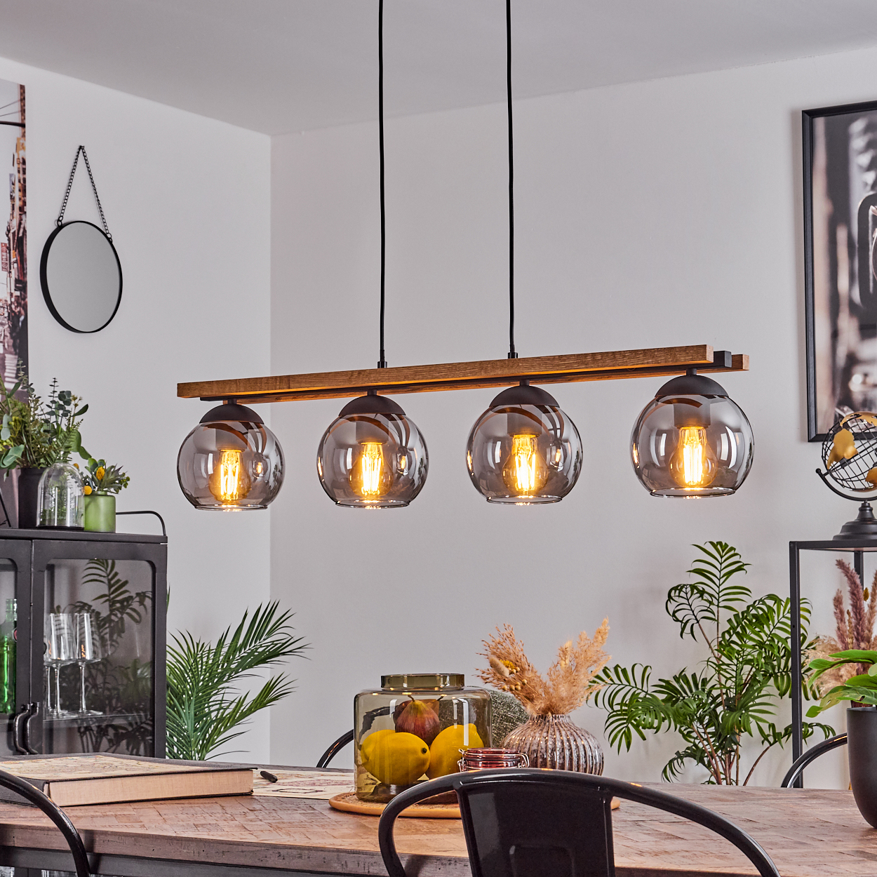 Esszimmer mit Holztisch, Pendelleuchten mit Glasschirmen, Pflanzen und einem Spiegel an der Wand. Sonnenlicht erhellt die Szene.