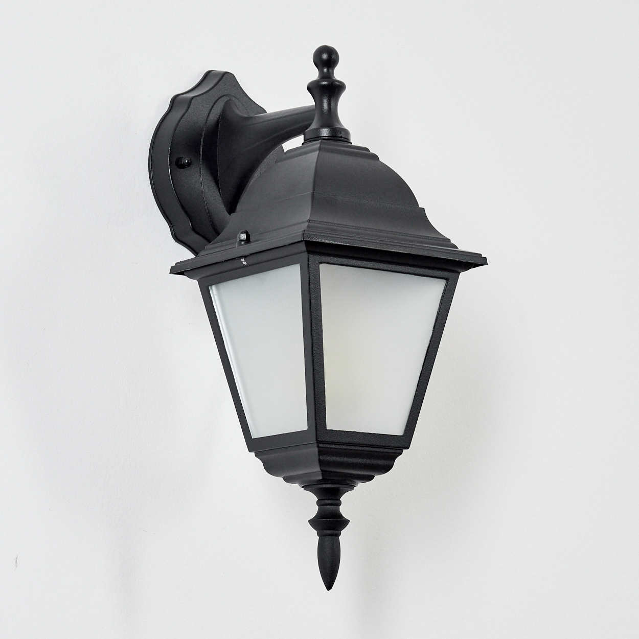 Eine schwarze Außenwandlaterne mit einer Milchglasscheibe ist an einer weißen Wand befestigt. Die Laterne hat ein dekoratives Design.