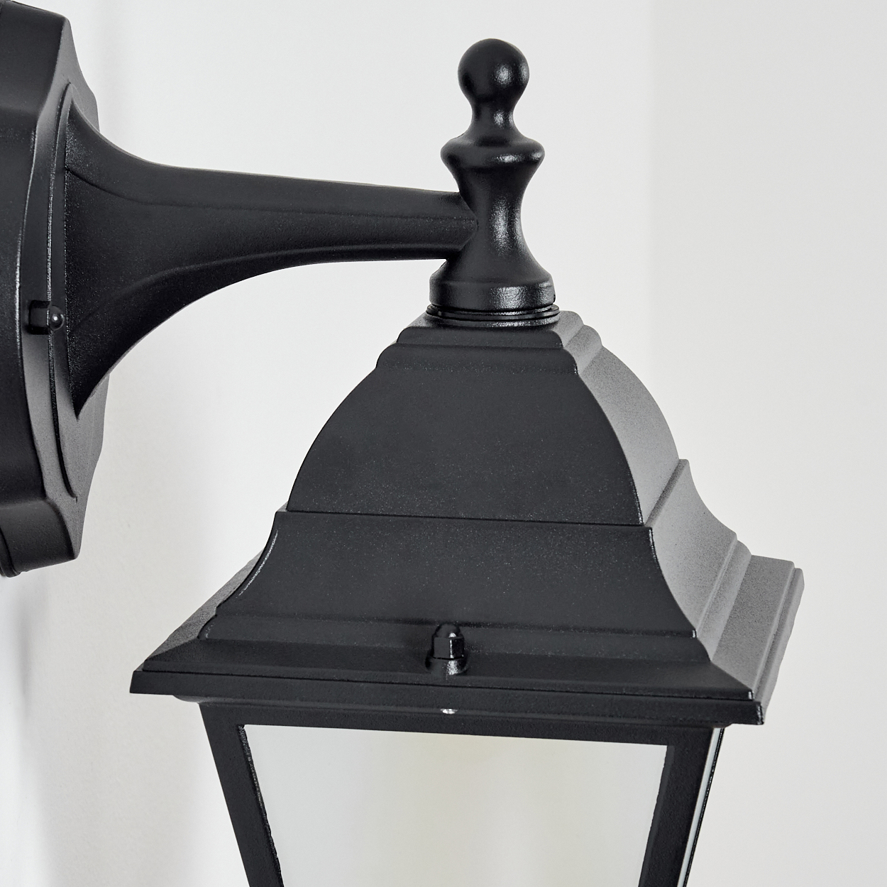 Schwarze Außenlampe an einer weißen Wand. Die Lampe hat ein Laternen-Design und eine dekorative obere Spitze.