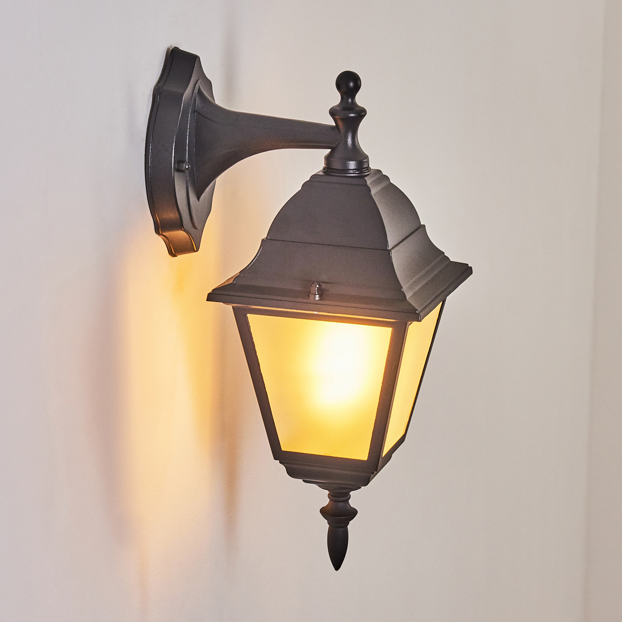 Eine schwarze Außenlaterne ist an einer weißen Wand montiert. Die Laterne ist beleuchtet und wirft einen warmen Schein. Die Szene ist gut beleuchtet.