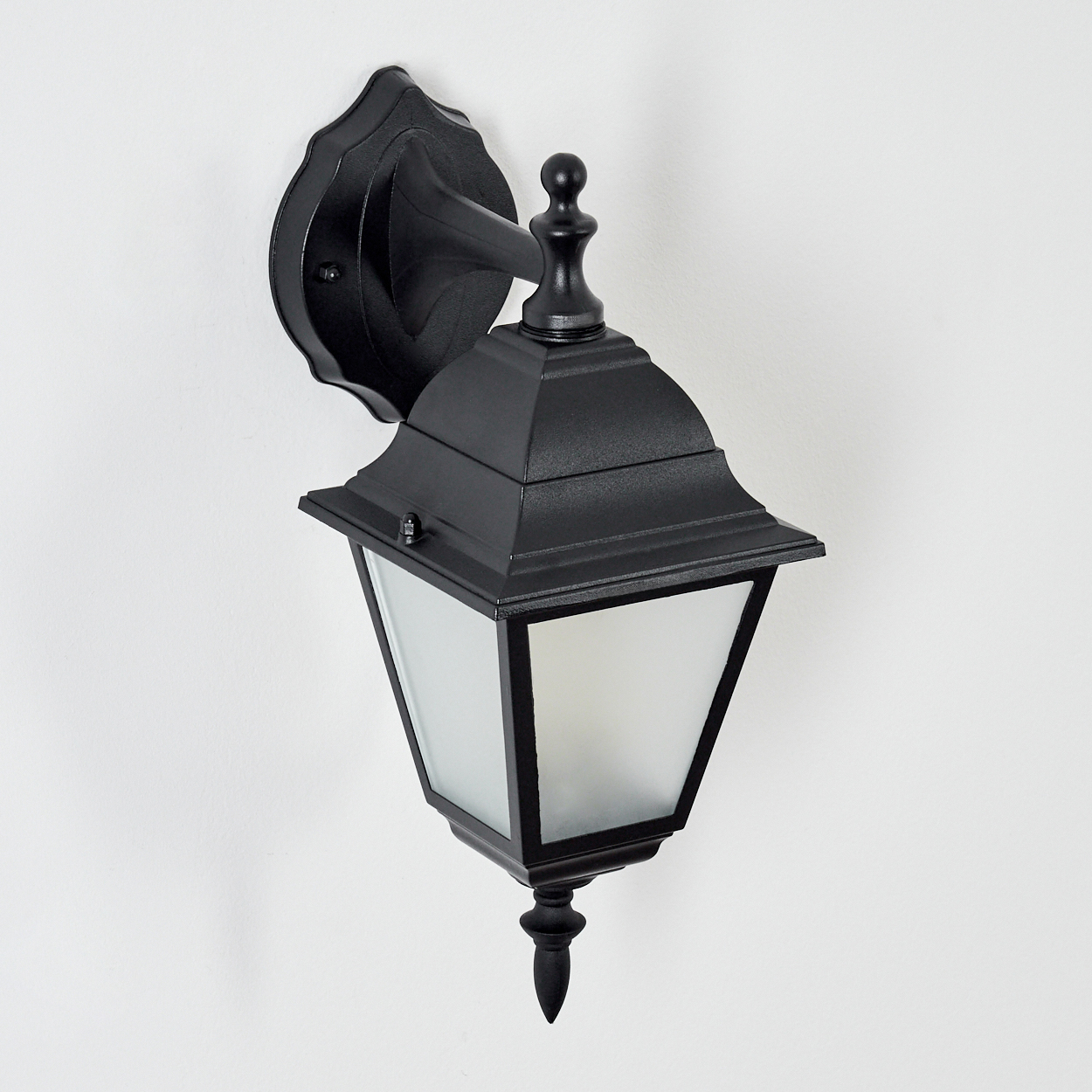 Eine schwarze Außenlampe ist an einer weißen Wand befestigt. Die Lampe hat eine Laternenform mit Milchglasscheiben und einem dekorativen Aufsatz.