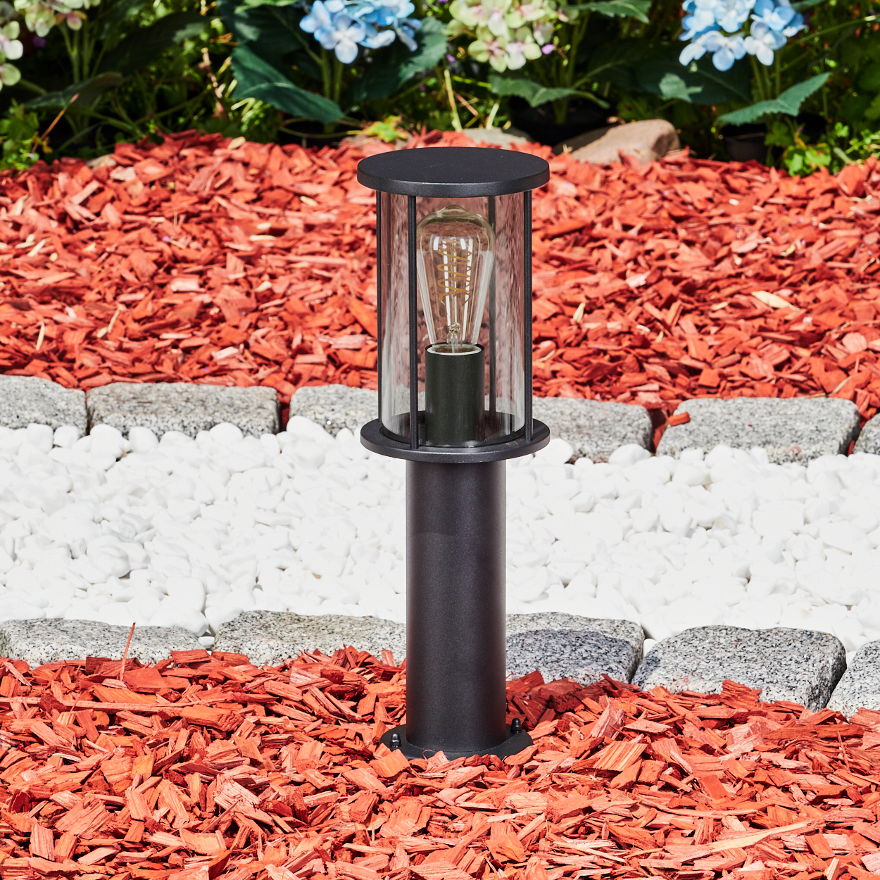 Eine schwarze Außenlampe mit Glasgehäuse und sichtbarer Glühbirne, eingebettet in ein Blumenbeet mit roter Mulche und weißen Steinen.