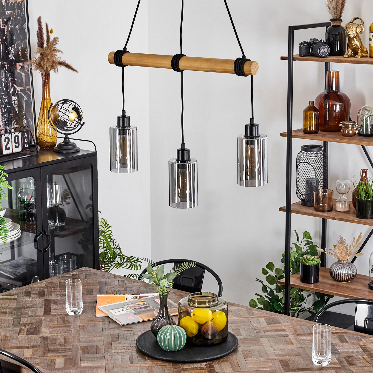 Esszimmer mit Holztisch. Drei Pendelleuchten hängen darüber. Ein Regal hält Dekorationen. Ein Glas mit Zitronen steht auf dem Tisch.