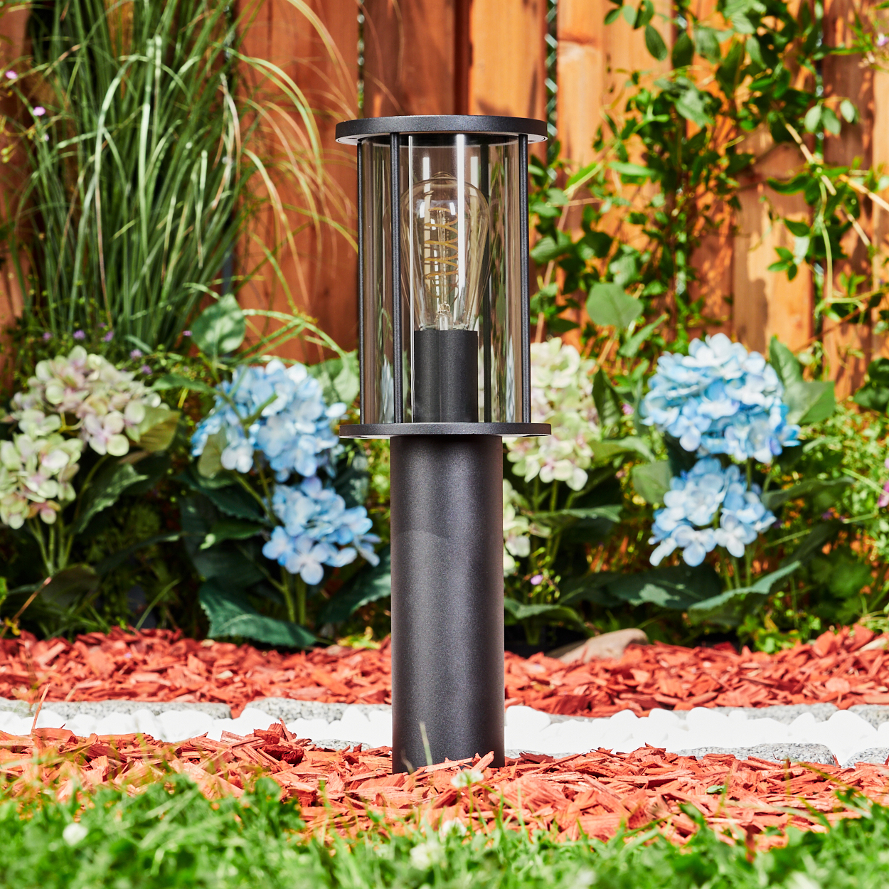 Eine schwarze Außenlampe mit sichtbarer Glühbirne steht in einem Garten. Blaue Hortensien und roter Mulch umgeben sie. Im Hintergrund ist ein Holzzaun zu sehen.