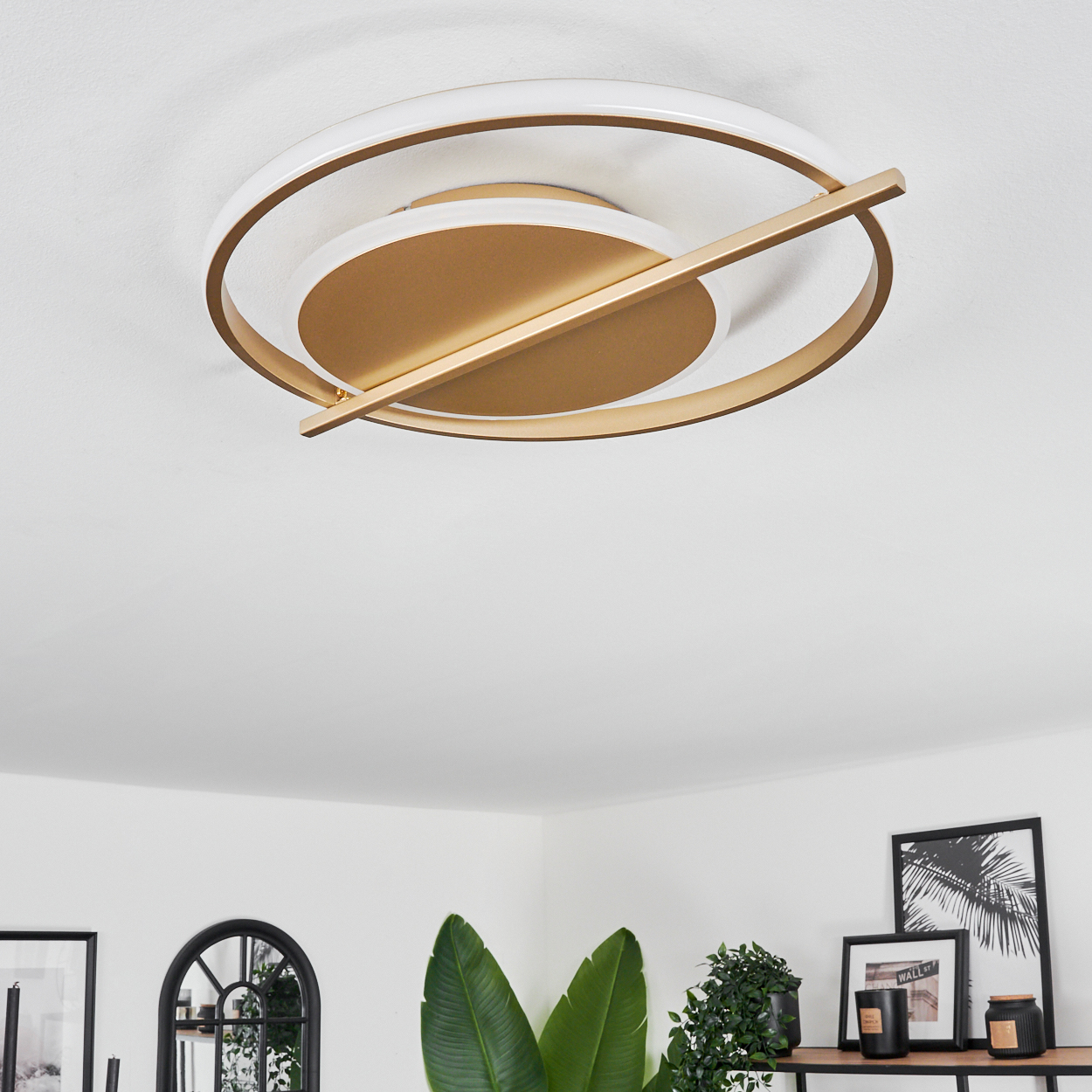 Eine Deckenleuchte mit goldenen und weißen Ringen. Ein goldener Balken kreuzt die Ringe. Darunter ein Regal mit Pflanzen, Kerzen und Bildern.