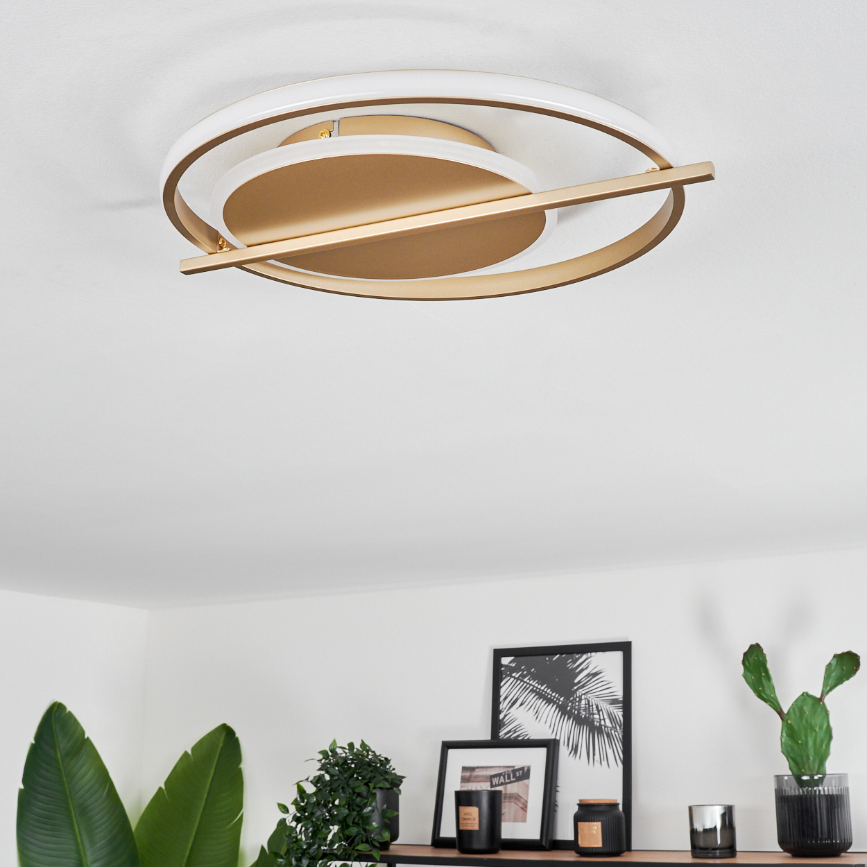 Moderne Deckenleuchte mit Gold- und Weißakzenten. Darunter ein Regal mit Pflanzen und gerahmter Kunst, darunter ein Palmen-Druck.