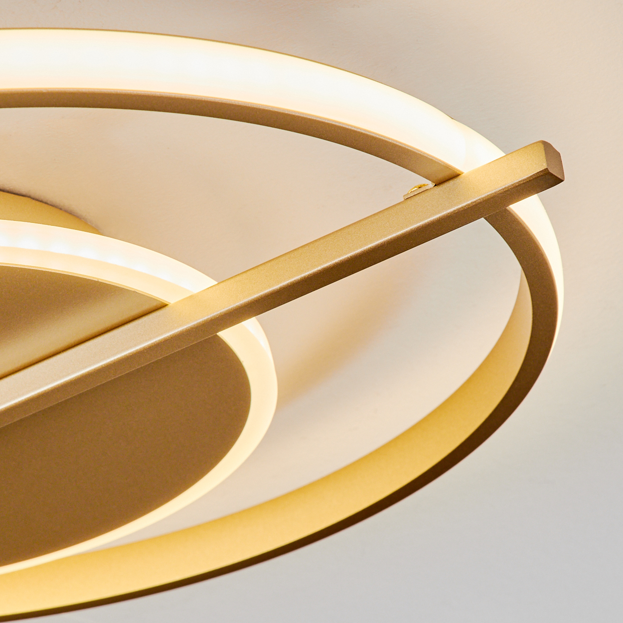 Nahaufnahme einer modernen Deckenleuchte. Goldfarbene Metallkreise und ein Balken schneiden sich, mit hellem, warmweißem Licht.