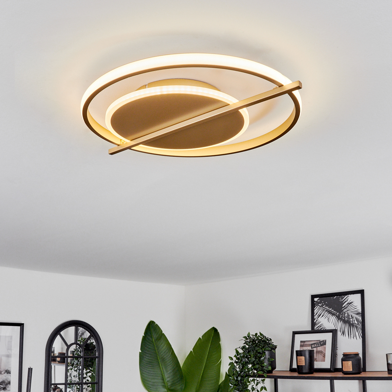 Moderne Deckenleuchte mit goldenen Ringen. Darunter Pflanzen, Bilder und Dekoration auf einem Regal vor einer wei ßen Wand.