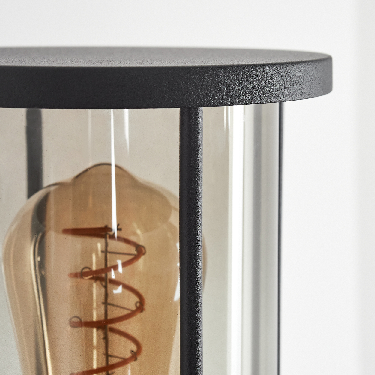 Nahaufnahme einer schwarzen Lampe mit Glasgehäuse. Innen ist eine große bernsteinfarbene Glühbirne mit rotem Faden sichtbar.