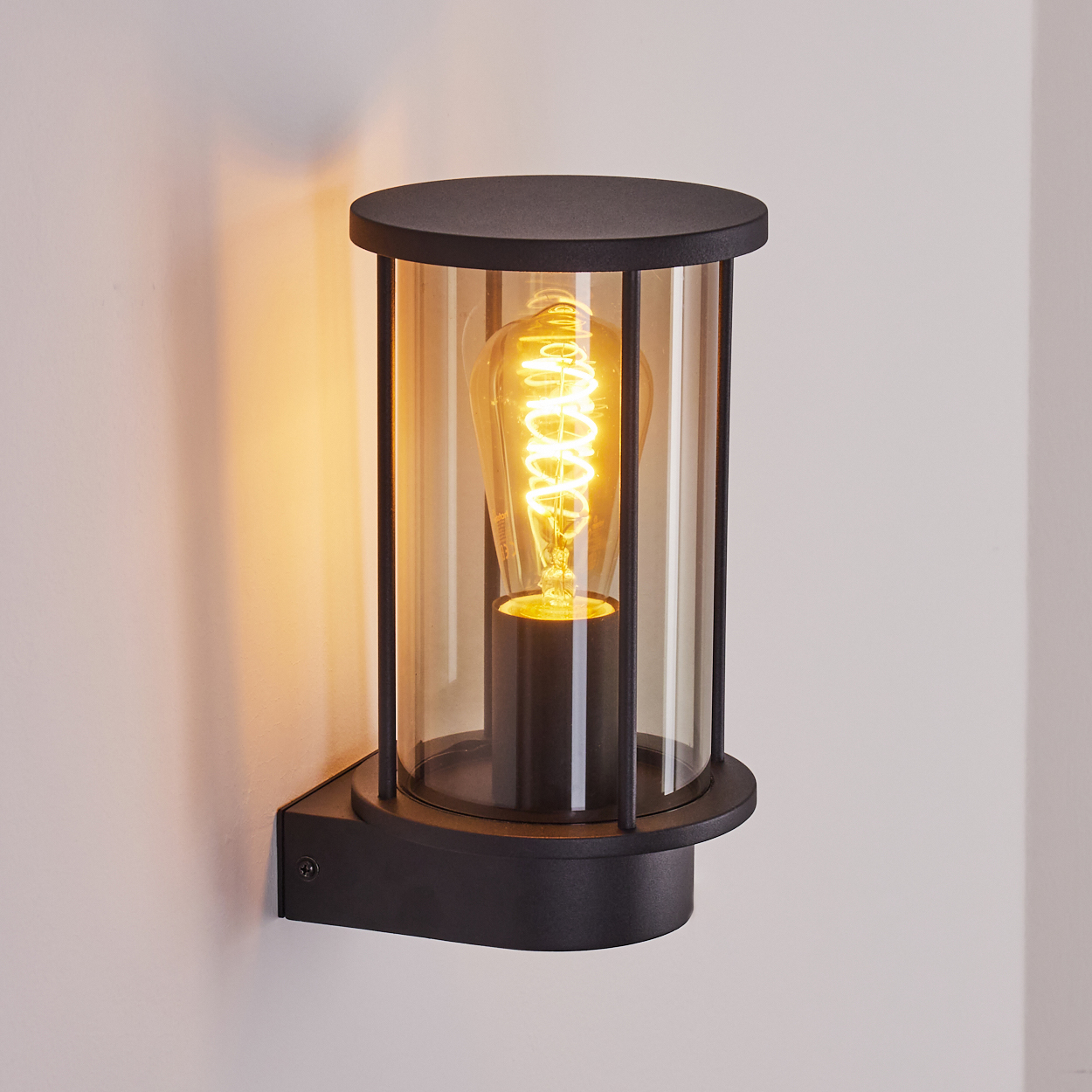 Eine schwarze Wandlampe mit zylindrischem Glasschirm und einer leuchtenden Wendelglühbirne. Sie ist an einer weißen Wand montiert.