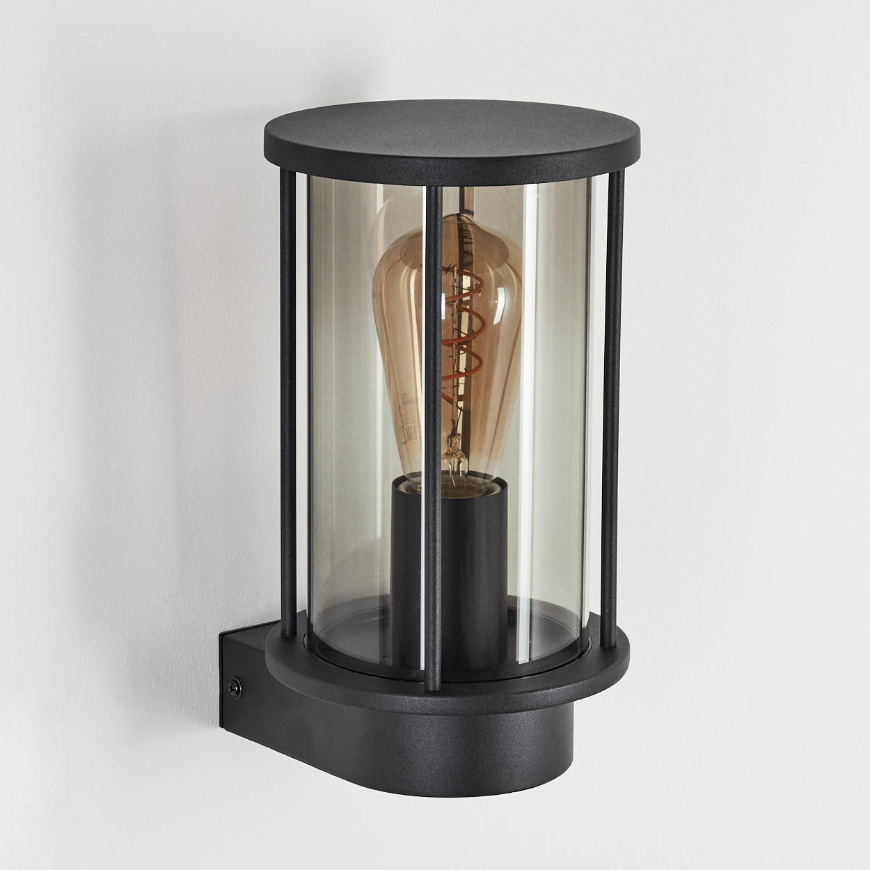 Schwarze Außenwandleuchte mit zylindrischem Glasschirm, der eine sichtbare Glühbirne beherbergt, an einer weißen Wand montiert.