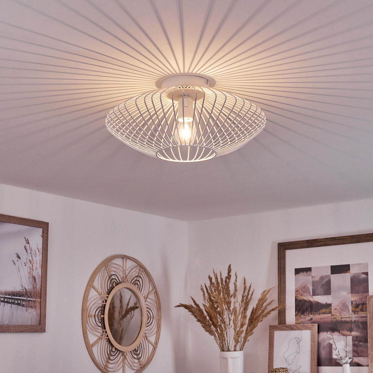 Eine weiße, moderne Deckenlampe wirft Schatten an die Decke. Darunter befinden sich ein Spiegel, gerahmte Kunst und Trockenpflanzen in einer Vase.