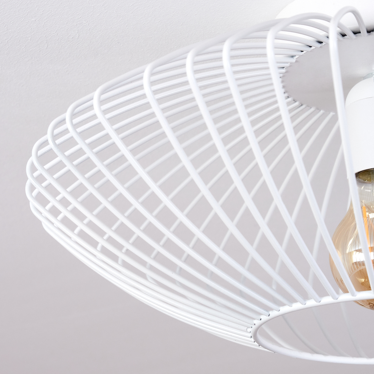 Eine weiße Deckenlampe mit Drahtrahmen und sichtbarer Glühbirne. Die Lampe ist an einer weißen Decke montiert. Der Stil ist modern.