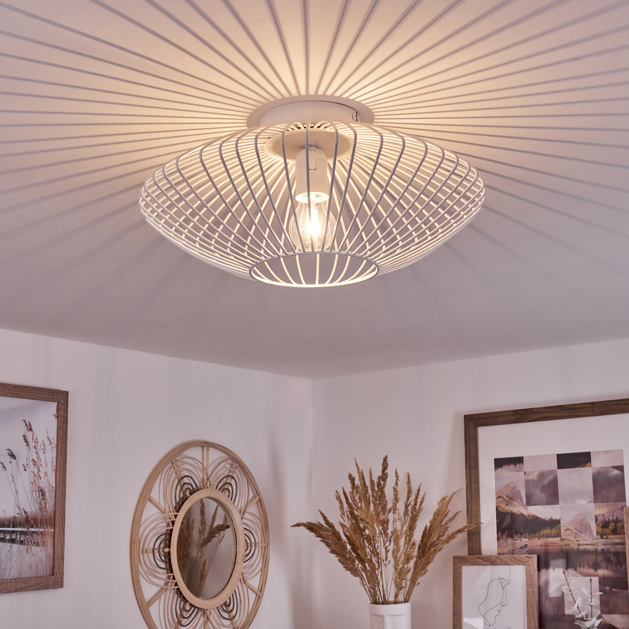 Eine weiße Deckenlampe mit Käfig wirft einen radialen Schatten auf die Decke. Darunter ein Raum mit Spiegel, Bildern und einer Vase mit getrockneten Pflanzen.