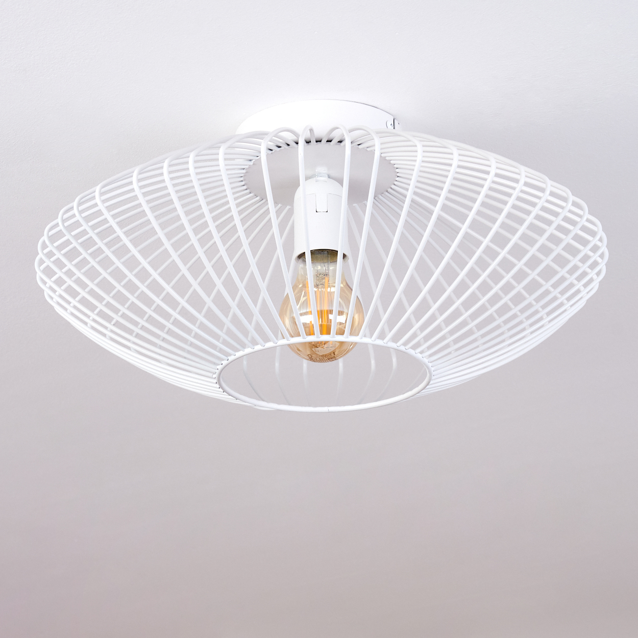 Weiße Deckenlampe mit Drahtrahmen und sichtbarer Glühbirne. Die Lampe ist vor einer weißen Decke zentriert. Die Glühbirne hat ein warmes Leuchten.