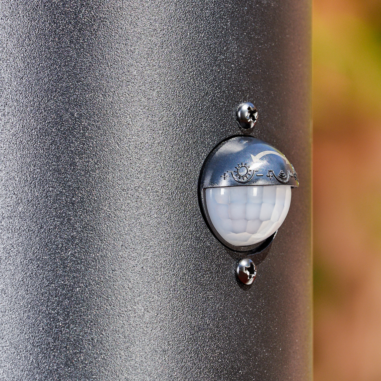 Nahaufnahme einer dunkelgrauen Metallstange mit einem Bewegungssensor. Der Sensor hat eine weiße, abgerundete Linse und ist mit Schrauben befestigt.
