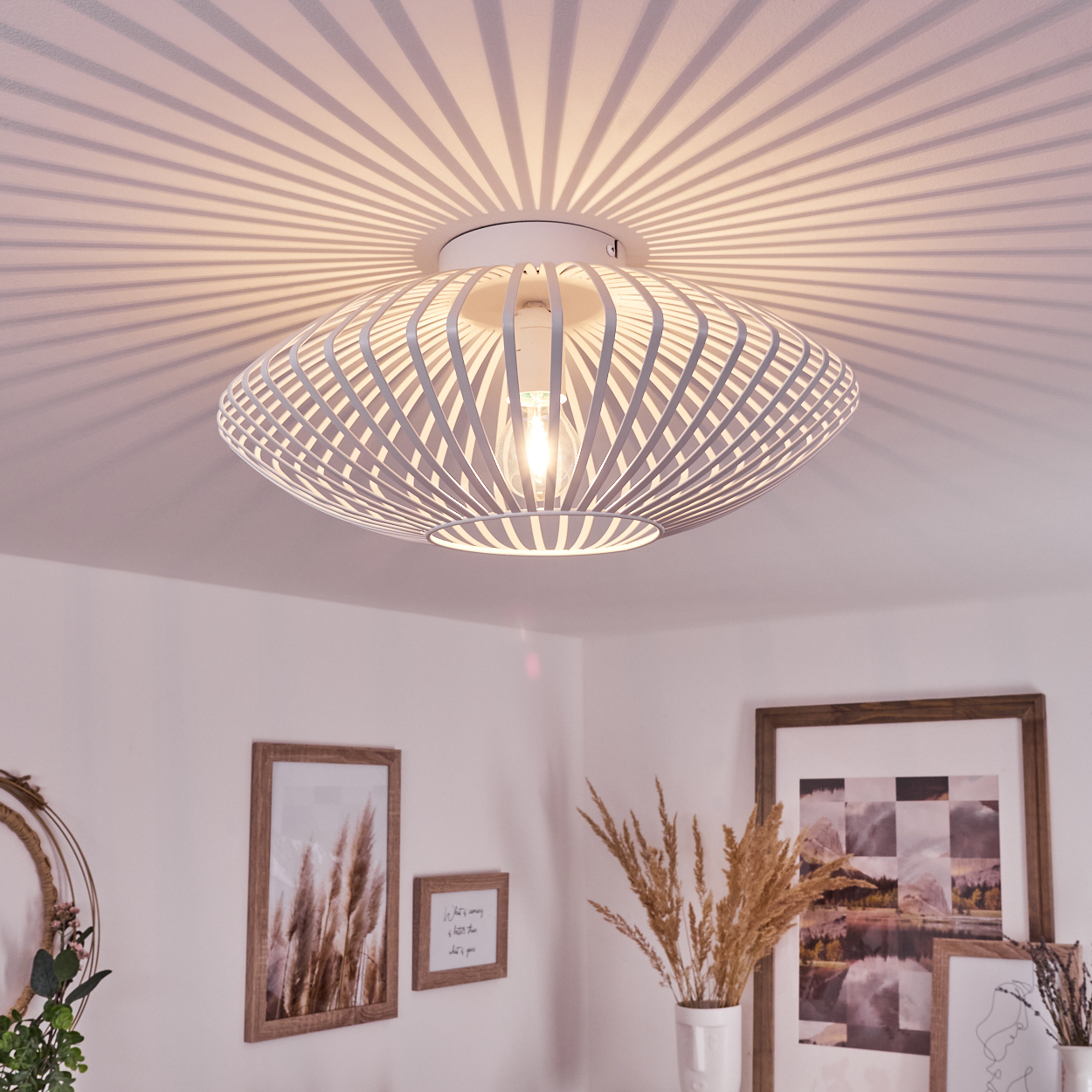 Eine weiße Deckenlampe mit einzigartigem Design wirft Schatten auf die Decke. Rahmen mit Bildern und Vasen mit Pflanzen befinden sich im Raum.