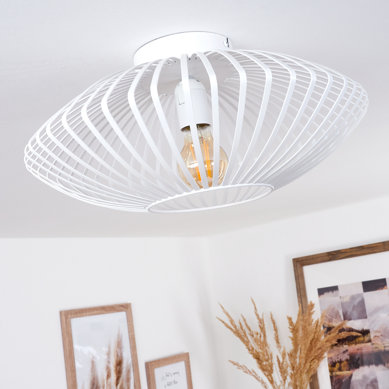 Eine weiße, ovale Deckenlampe mit sichtbarer Glühbirne. Darunter sind gerahmte Bilder und getrocknete Pflanzen zu sehen.