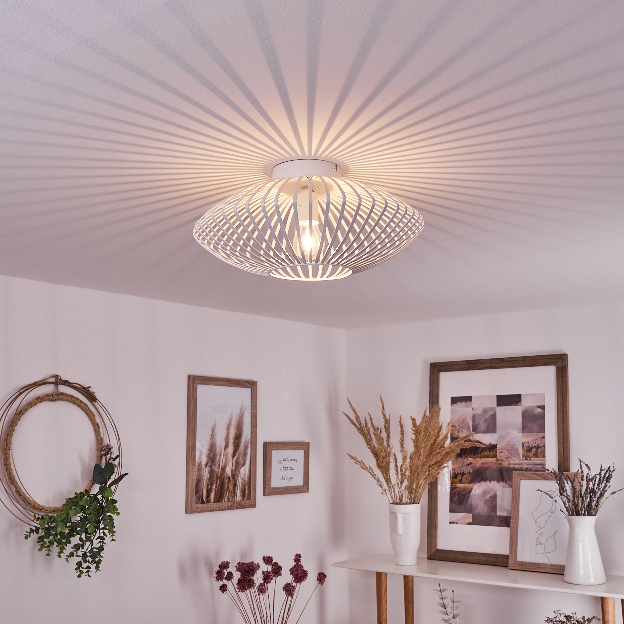 Eine weiße, moderne Deckenlampe wirft Licht und Schatten. Der Raum hat gerahmte Bilder, Vasen mit Pflanzen und einen Blumenschmuck.