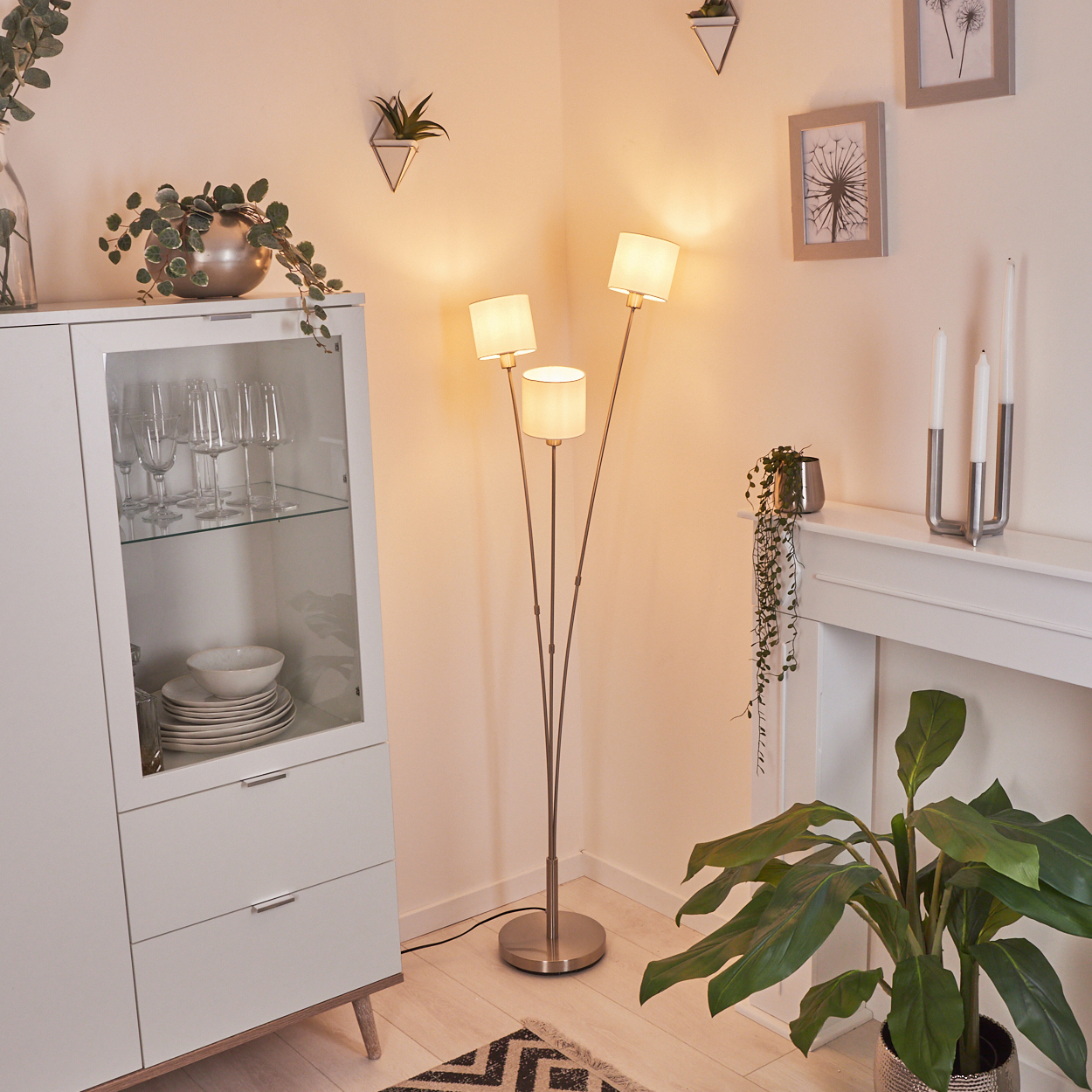 Ein Raum mit einem weißen Schrank, einer Stehlampe und einer Pflanze. An der Wand befinden sich dekorative Rahmen und eine kleine Hängepflanze.